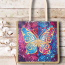 A canvas tote bag with a colorful printed design featuring a large glittery butterfly over a mandala pattern in blue, pink, purple, and gold. The bag has natural beige handles and a beige border, shown on a rustic white wood background with cotton bolls as a decorative prop.