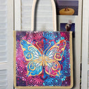A canvas tote bag with cream-colored shoulder straps, featuring a large, vibrant, rhinestone-embellished butterfly design on a purple and pink mandala background. The bag is displayed hanging against a white wooden shutter door with a small picture frame clipped to the side.