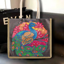 A tote bag with a colorful peacock and floral design, set against a dark background with a black bag partially visible behind it.