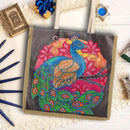 A canvas tote bag with a colorful beaded peacock design, styled on a white lace surface with decorative props including a small book, blue pencils, and a gift box.