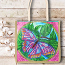 A canvas tote bag with cream-colored handles, featuring a colorful beaded-style butterfly and leaf design on a pink background, styled on a rustic white wood surface next to cotton bolls.