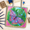 A diamond painting or bead art kit canvas featuring a colorful butterfly on green leaves, surrounded by pink and blue decorative elements on a white wooden background.