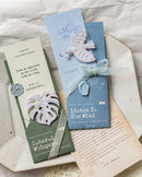 Two decorative paper bookmarks or card bookmarks styled on a ceramic dish over a cream fabric background. One bookmark is sage green with a white monstera leaf and the text 'Natural Whisper'.