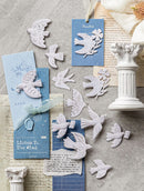 A styled flat-lay of white paper cutout birds and matching blue greeting cards with printed text, arranged on a gray stone-like surface with vintage paper, a white column prop, and a cream ribbon.