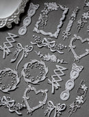 A collection of white embossed paper or cardstock embellishments with ornate floral frames, bows, ribbons, and decorative motifs arranged on a dark gray textured background.