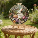 A circular embroidered artwork featuring a white bunny with an orange egg and colorful flowers, displayed on a small wooden stand atop a woven wicker stool in a garden setting.