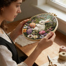 A woman holds a round embroidery hoop featuring a detailed embroidered scene of a white rabbit among colorful flowers and Easter eggs. The setting is a warm, softly lit craft room with a wooden table, embroidery floss, and scissors in the background.