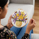 A woman holds a circular embroidery hoop featuring a colorful floral bouquet with yellow flowers, purple blossoms, butterflies, and a woven basket. She is shown in profile, sewing in a real indoor setting with a light gray sofa and wooden floor visible.