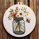 A circular embroidery hoop mounted on a rustic wooden wall, featuring a floral bouquet in a glass jar with a bee beside it. The embroidery includes white daisies, pink and yellow flowers, green leaves, and black-and-yellow stripes on the bee.