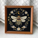 A framed square art piece featuring a detailed embroidered or printed bee with white wings, surrounded by white flowers, small stars, and a gold crescent moon on a black background, set in a medium brown wooden frame.