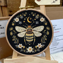 A round embroidery hoop displaying a finished black embroidered design of a bumblebee, white flowers, a crescent moon, and stars, shown on a small wooden stand.