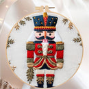 A close-up of a circular embroidery hoop holding white fabric with a detailed embroidered nutcracker figure in red, navy, white, and gold, surrounded by small gold leaf motifs.