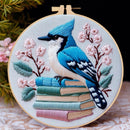 A round embroidery hoop displays a detailed bluebird perched on a stack of books, surrounded by pink blossoms and green leaves. The hoop is set against a soft-focus background with green foliage and a dark upper area.