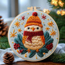 A circular embroidery hoop featuring a festive embroidered bird wearing a yellow-orange knit hat and red scarf, surrounded by green pine branches, red berries, and gold star motifs on a light blue fabric background. The hoop is styled on a wooden surface with pinecones, a knit cloth, and blurred holiday lights in the background.