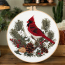 A circular embroidery hoop displays a detailed needlepoint or embroidery design of a red cardinal perched on snowy pine branches with pinecones and holly leaves. The hoop is shown resting on a wooden surface with a softly blurred home decor background.
