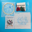 Embroidery DIY art kit laid out on a bright blue background, including an embroidery hoop with thread, a package of multicolor embroidery floss, a pattern booklet showing a cardinal design, and a white fabric square with a blue cardinal embroidery outline.