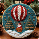 A circular embroidery hoop displays a festive Christmas-themed stitched design featuring a red and white striped hot air balloon with Santa Claus in a basket, flanked by two green Christmas trees on a teal background with white snowy ground. The hoop is styled with pine branches, pinecones, and warm lights in the background.
