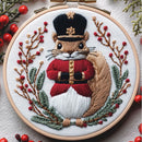 A circular embroidery hoop holding a detailed embroidered nutcracker squirrel design. The squirrel wears a black top hat with a gold emblem, a red military-style coat with gold buttons, and a white belly, surrounded by green pine branches and red berries on a white fabric background.