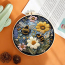 A circular embroidery hoop displaying a detailed floral and bee design, styled on an orange surface with a book, succulent, and pinecones.