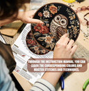 Person holding a round embroidered piece with an owl design, surrounded by crafting materials and an instruction manual.