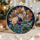 A round embroidery hoop containing a colorful peacock design with fanned feathers, photographed in a softly lit indoor setting with blurred holiday lights and ornaments in the background.