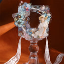A decorative clear acrylic picture frame with a floral and butterfly embellishment border, a printed illustration of a woman in a white dress, and white pearls, displayed on a clear glass pedestal against a warm brown background.