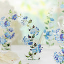 Blue and purple flowers with green leaves arranged on a white surface against a soft, bright, blurred background.