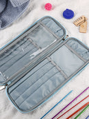 An open light blue and gray knitting needle case with multiple compartments, shown on a white textured blanket with knitting accessories like colorful needles, a blue cable, and wooden tools.