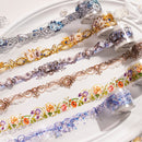 A close-up of multiple rolls of decorative washi tape laid out on a white surface. The tapes feature ornate lace-like patterns and floral motifs in silver, gold, bronze, blue, purple, and multicolor designs.