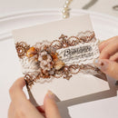 A close-up of a hand holding a decorative card with ornate lace, 3D floral embellishments, and the word 'Romance' printed on it. Another hand is visible at the right edge against a soft white background.