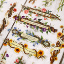 A flat-lay view of decorative paper or cardstock featuring vintage-style botanical illustrations, including sunflowers, roses, purple flowers, and twisted vines with printed inspirational quotes on a distressed white wood background.