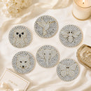 Six round beaded coasters featuring animal and nature designs including an owl, hummingbird, bee, fox, elephant, and butterfly, arranged on a cream satin fabric with decorative props.