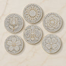 Six round gold-toned brooches embellished with white stones in floral and geometric patterns, arranged on a light beige marble surface.