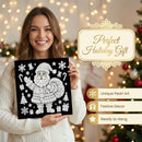 A smiling woman in a cream knit sweater holds up a black square canvas featuring a white illustrated Santa Claus surrounded by holiday motifs. The background is softly blurred with warm string lights and a Christmas tree, and decorative text on the right reads 'Perfect Holiday Gift' with feature callouts.