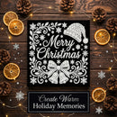 A square black Christmas-themed plaque or sign with white ornate holiday illustrations and the words “Merry Christmas,” styled on a dark wood surface with dried orange slices, pinecones, fairy lights, and silver snowflake decorations. A lower banner reads “Create Warm Holiday Memories.