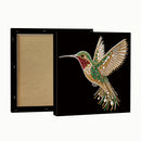 A square black canvas or framed print shown from the back and side, featuring a jeweled hummingbird design in gold, green, red, and white on a black background. The left side shows the plain tan backing board.