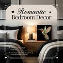 A framed decorative wall art piece featuring a colorful hummingbird on a black background, styled in a bedroom setting beside a lamp and between two beds. Large overlaid text reads 'Romantic Bedroom Decor' with smaller descriptive copy.