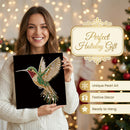 A woman in a white knit sweater holds a black square panel featuring a colorful hummingbird made of rhinestones on a black background. The scene is festive with warm holiday lights and a decorated Christmas tree behind her, and text on the right reads 'Perfect Holiday Gift' with additional product callouts.