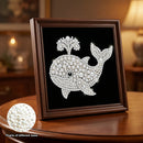 A framed whale-shaped pearl and crystal artwork displayed on a wooden table in a warmly lit room. The whale design is made of white pearls and rhinestones on a black background, inside a dark brown wooden frame.