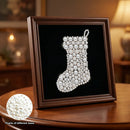 A square framed Christmas stocking craft piece made of many pearl-like beads in different sizes on a black background, displayed on a wooden table in a warmly lit room. A circular inset shows a close-up of the pearl texture.