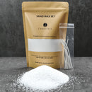 A sand wax set kit by Candittle, featuring a gold pouch, a small pile of white wax powder, and a clear plastic bag containing wicks, arranged on a dark gray surface against a textured gray background.