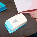 A small craft die-cut paper puncher with a cream top and light blue base, shown on a dark wooden table with a hand nearby and paper in the background.
