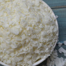 White wax flakes or soap shavings piled in a white bowl on a blue wooden surface.