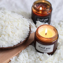 Two amber glass jar candles with white labels reading 'thank you HAND MADE' are styled on a white fabric background with scattered white wax flakes. One candle is lit in the foreground, and another is lit in the background.