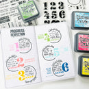 An open planner-style notebook with colorful motivational quote stickers and black ink stamps, surrounded by Distress Oxide ink pads and numbered stamp blocks on a white background.