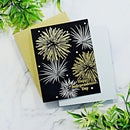 A handmade Independence Day greeting card with a black background and gold and white fireworks, layered over a gold envelope and a light blue card, styled on a white marble surface with green leaves.