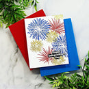 A white Independence Day greeting card with stamped red, blue, and gold fireworks, layered over red and blue envelopes on a white marble surface with green foliage.