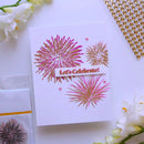 A white handmade greeting card featuring metallic pink and gold stamped fireworks and a banner that reads 'Let's Celebrate!', styled on a white surface with decorative flowers, gold beads, and a clear stamp packet.