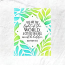A square art print featuring a Bible verse from Matthew 5:14 in black script and serif text, surrounded by a white scalloped border and framed by a watercolor leaf pattern in green, yellow, and blue. The print is displayed flat on a textured white surface.