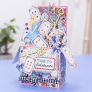 A 3D pop-up cardboard gift box featuring Alice in Wonderland themed illustrations, including a rabbit, Queen of Hearts, playing cards, a key, and the text 'TIME TO Celebrate'. The box is decorated with purple bows, a black and white checkered ribbon, and gold foil accents, set on a light blue surface with a blurred indoor background.